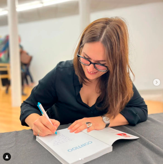 Mamen Jiménez, con un vestido negro, sentada detrás de una mesa cubierta de una tela gris, firma un ejemplar de su último libro llamado Contigo. Se la ve sonriente.