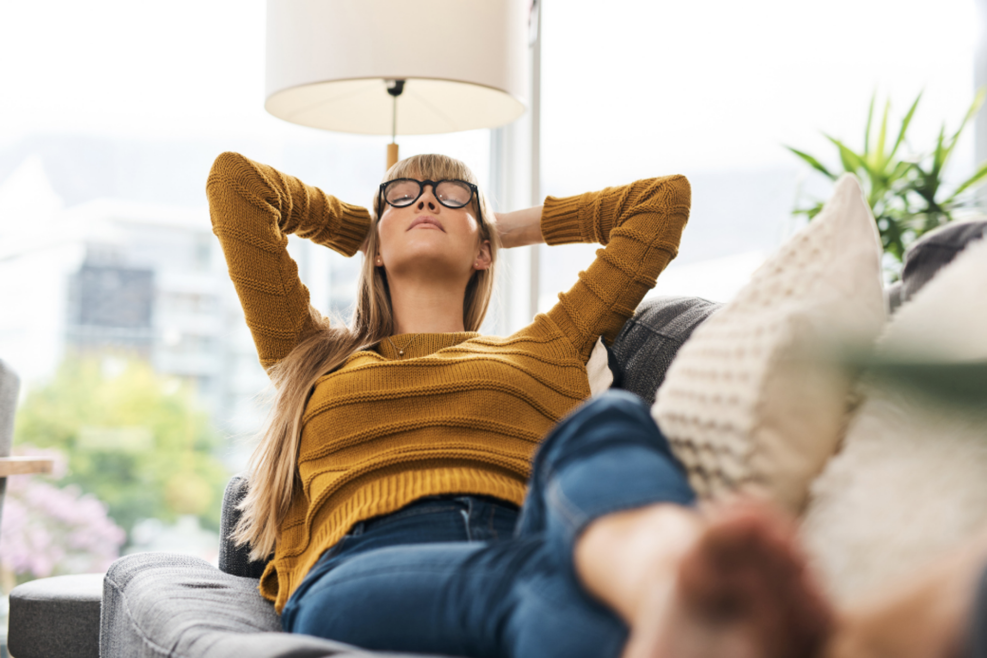 Mujer con gafas tumbada en el sofá, con las manos en la cabeza, sujetándose el cuello y con los ojos cerrados.