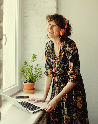 Chica con vestido de flores y auriculares puestos, sonríe ante su portátil, que está situado delante de una ventana.